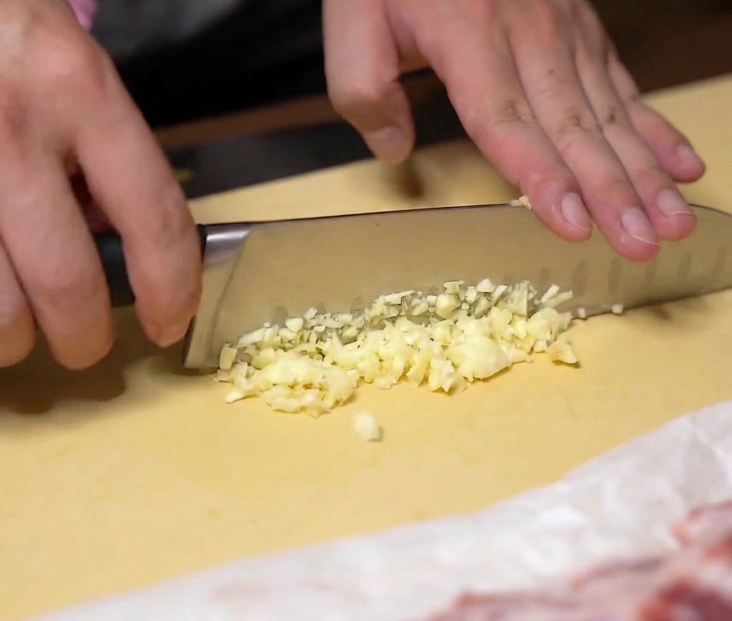 Hands finely mincing fresh garlic cloves with a large chefs knife on a yellow cutting board.