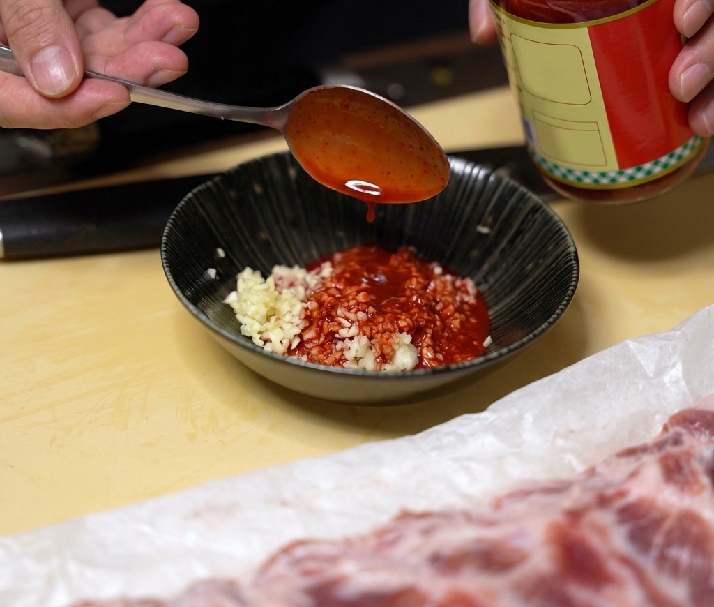 A spoonful of bright red fermented bean curd liquid being added to a black bowl containing minced garlic.