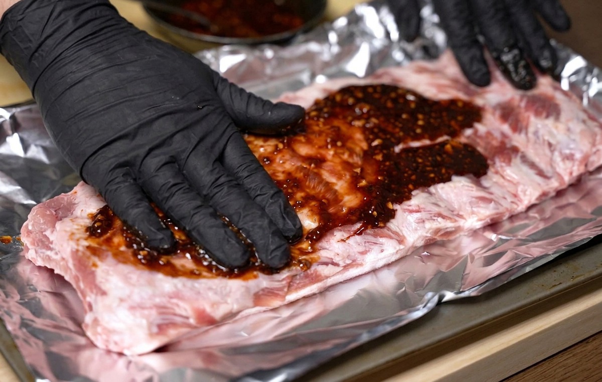 Gloved hands rubbing a thick, dark red marinade evenly over a large raw rack of pork ribs on aluminum foil.