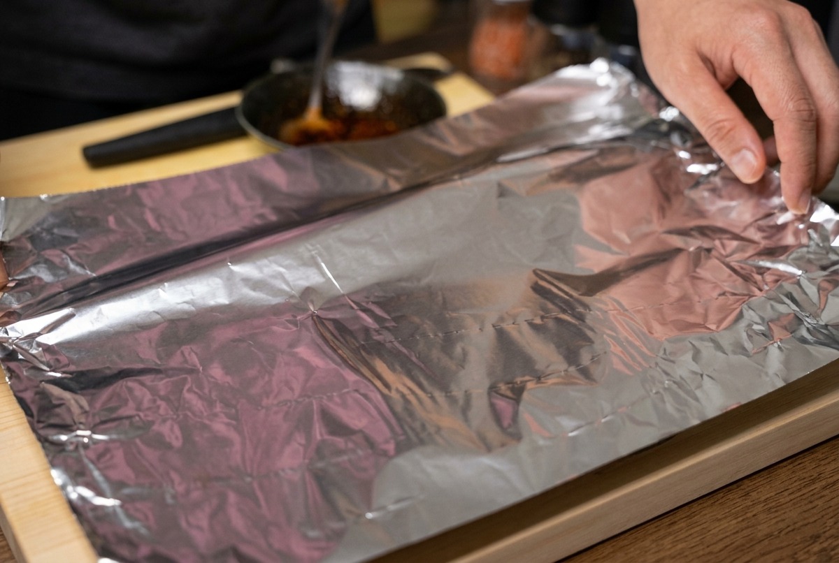 Hands folding a large sheet of aluminum foil to tightly wrap a rack of marinated pork ribs on a wooden board.