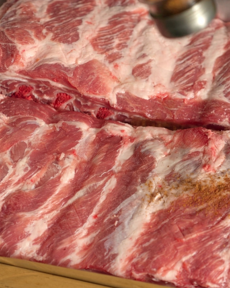 Dry spices being sprinkled from a shaker onto the surface of raw pork ribs.