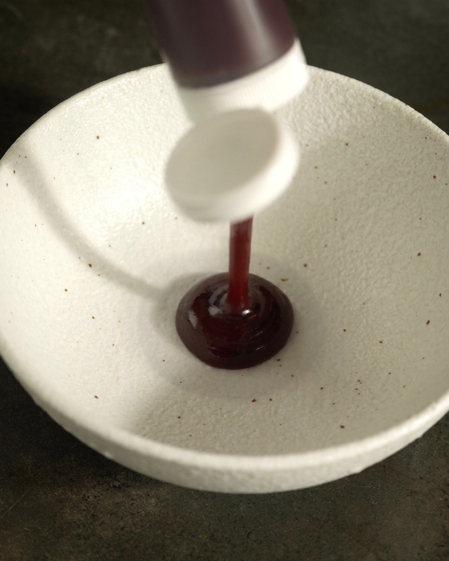 Thick, dark BBQ sauce being squeezed from a bottle into a white ceramic mixing bowl.