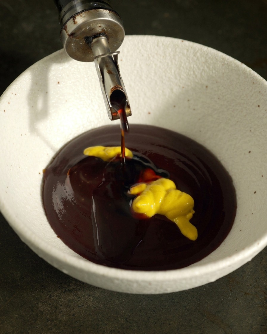 Light soy sauce pouring from a dispenser into a white bowl with dark BBQ sauce and a swirl of yellow mustard.