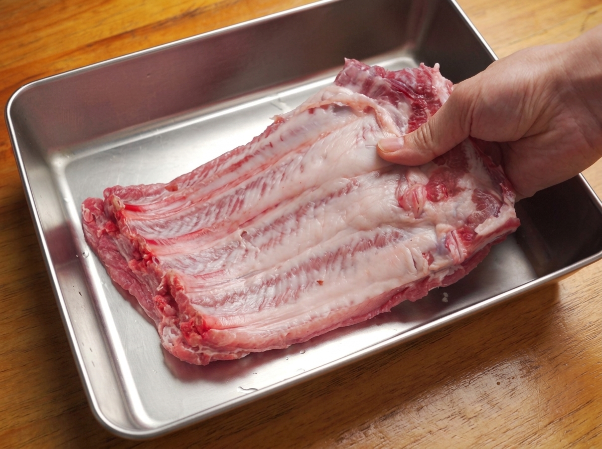 Hands pulling the thin membrane off the back of a large raw pork rib rack in a metal tray.