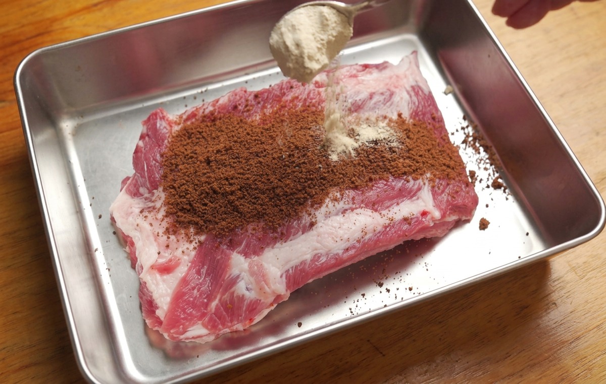 A tray holding raw pork ribs being sprinkled with brown sugar and a blend of dry spices from a spoon.