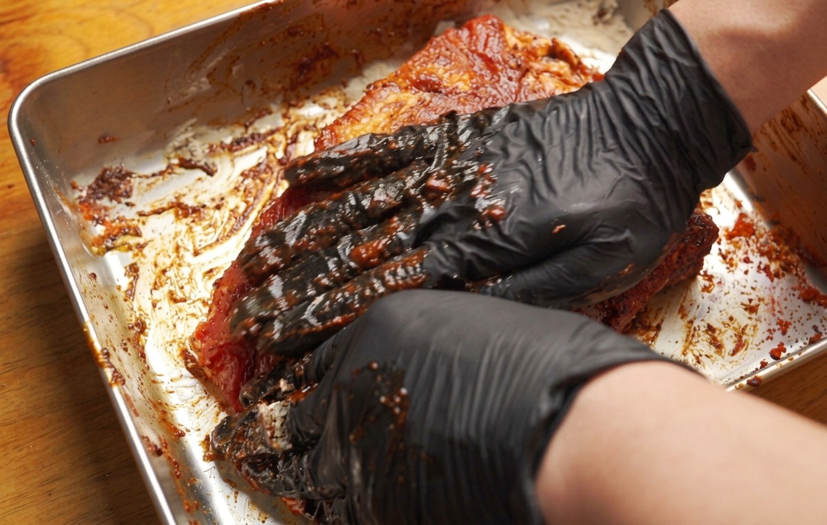 Gloved hands firmly rubbing a thick, reddish-brown marinade paste into a large slab of pork ribs in a metal tray.
