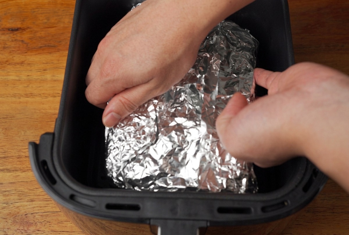 Hands placing a silver foil-wrapped package containing pork ribs into the black basket of an air fryer.