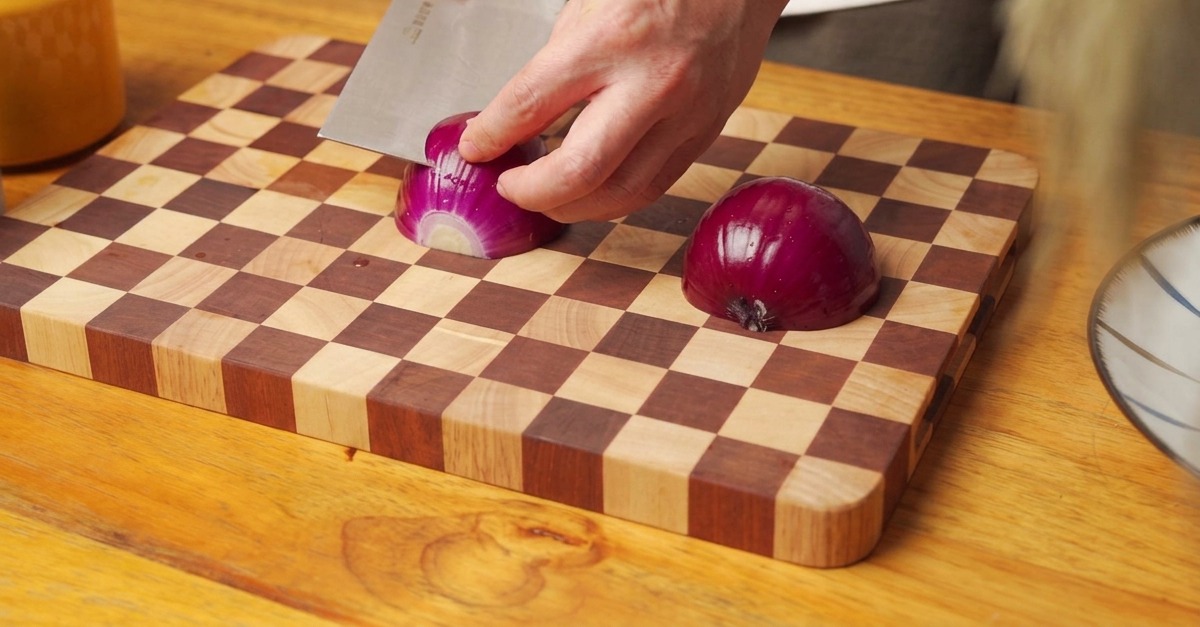 Hands using a cleaver to dice a red onion half on a wooden checkered cutting board.