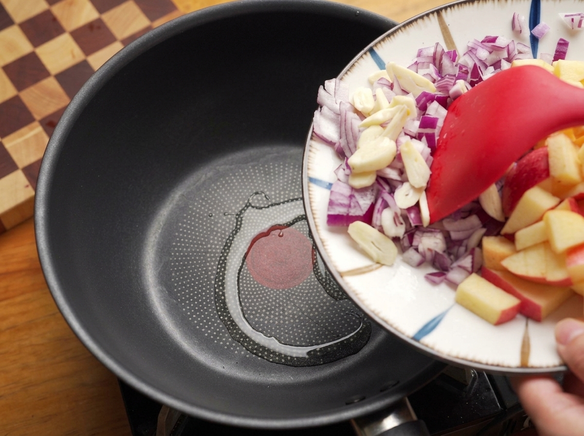 Using a spatula to slide chopped red onions and garlic from a plate into a heated pan with oil.