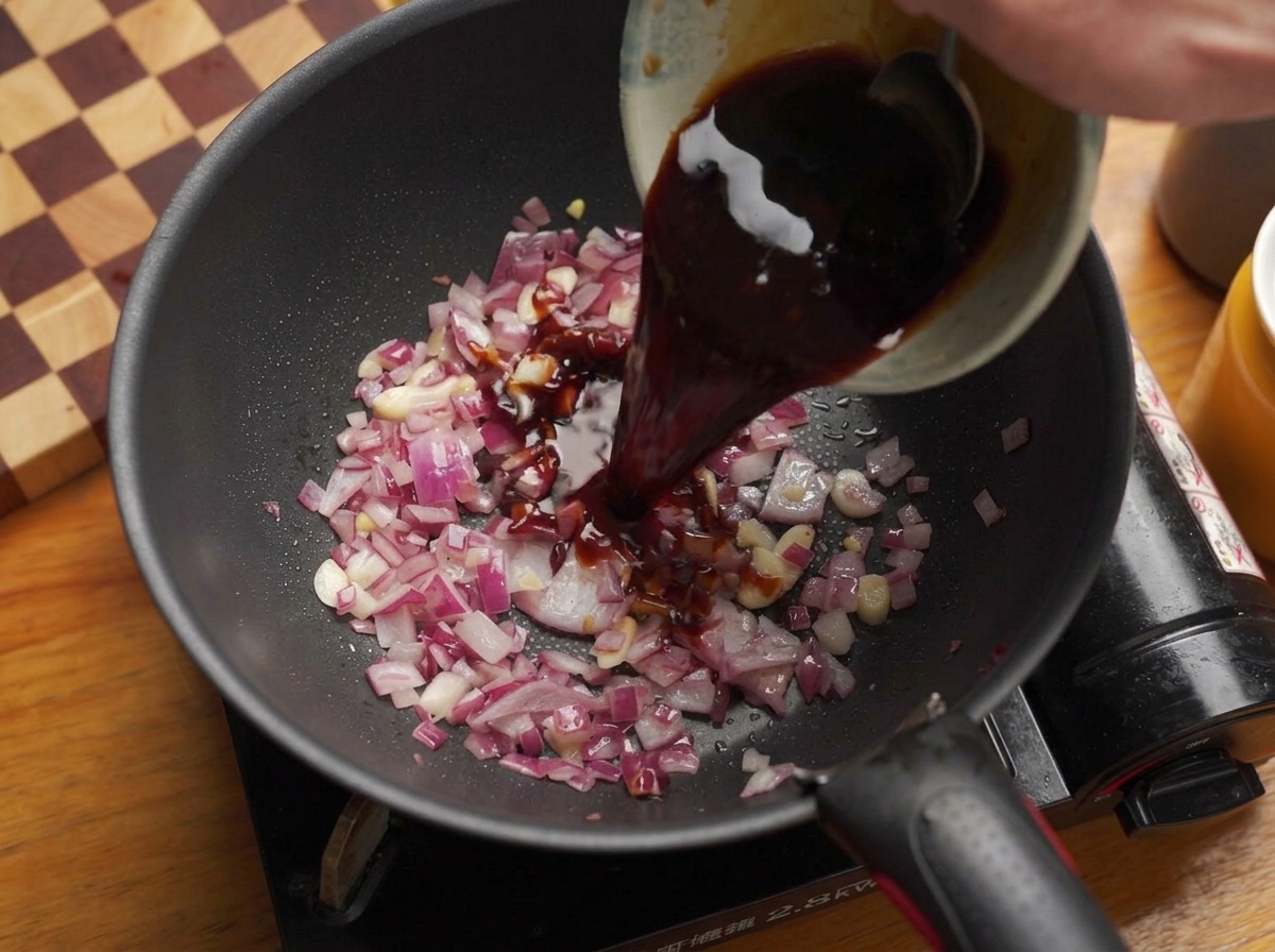 Pouring a dark, savory liquid sauce mixture from a bowl into a pan of sautéed onions and garlic.