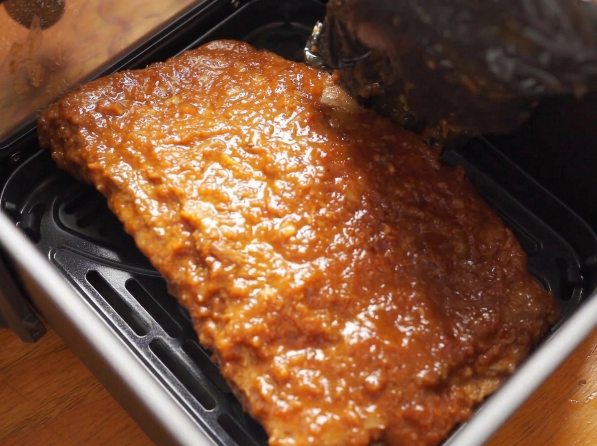 A black-gloved hand placing a large piece of pork ribs, thickly coated in a glossy barbecue sauce, down into an air fryer basket.