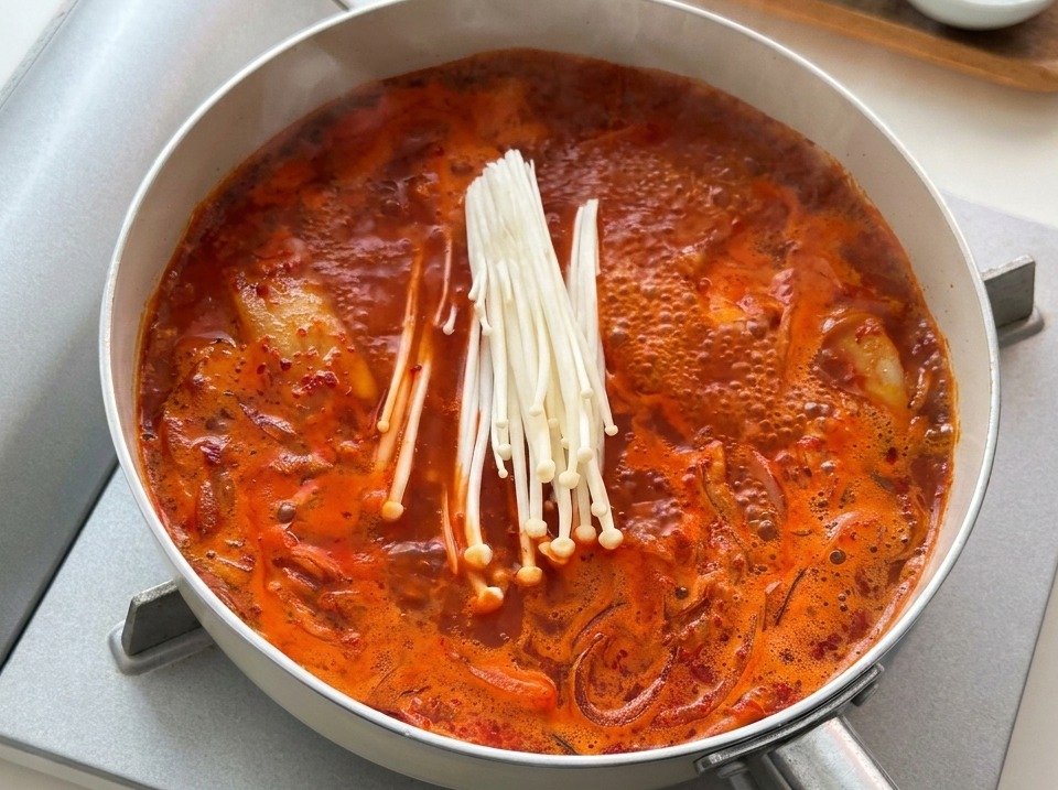 A bundle of raw white enoki mushrooms being added to a pan of boiling red kimchi stew.