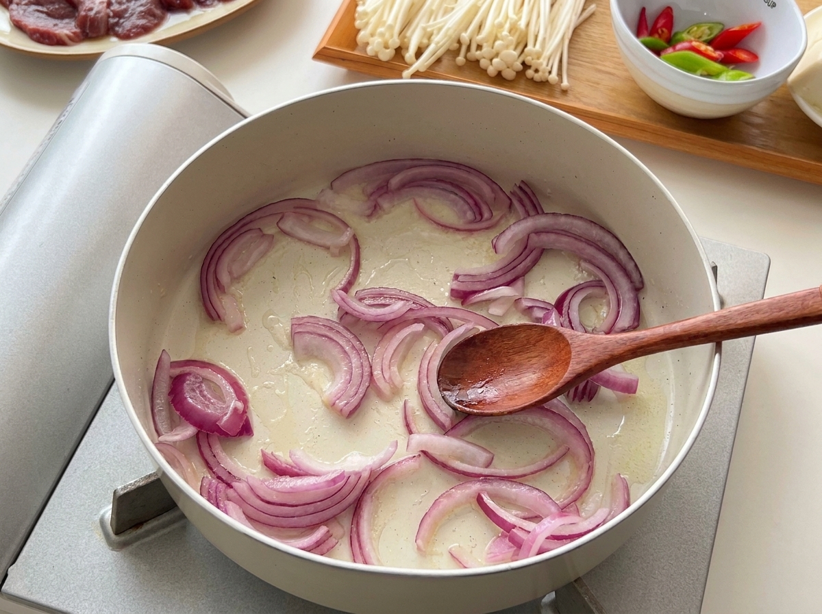 Sliced red onions being sautéed in a lightly oiled pan with a wooden spoon.