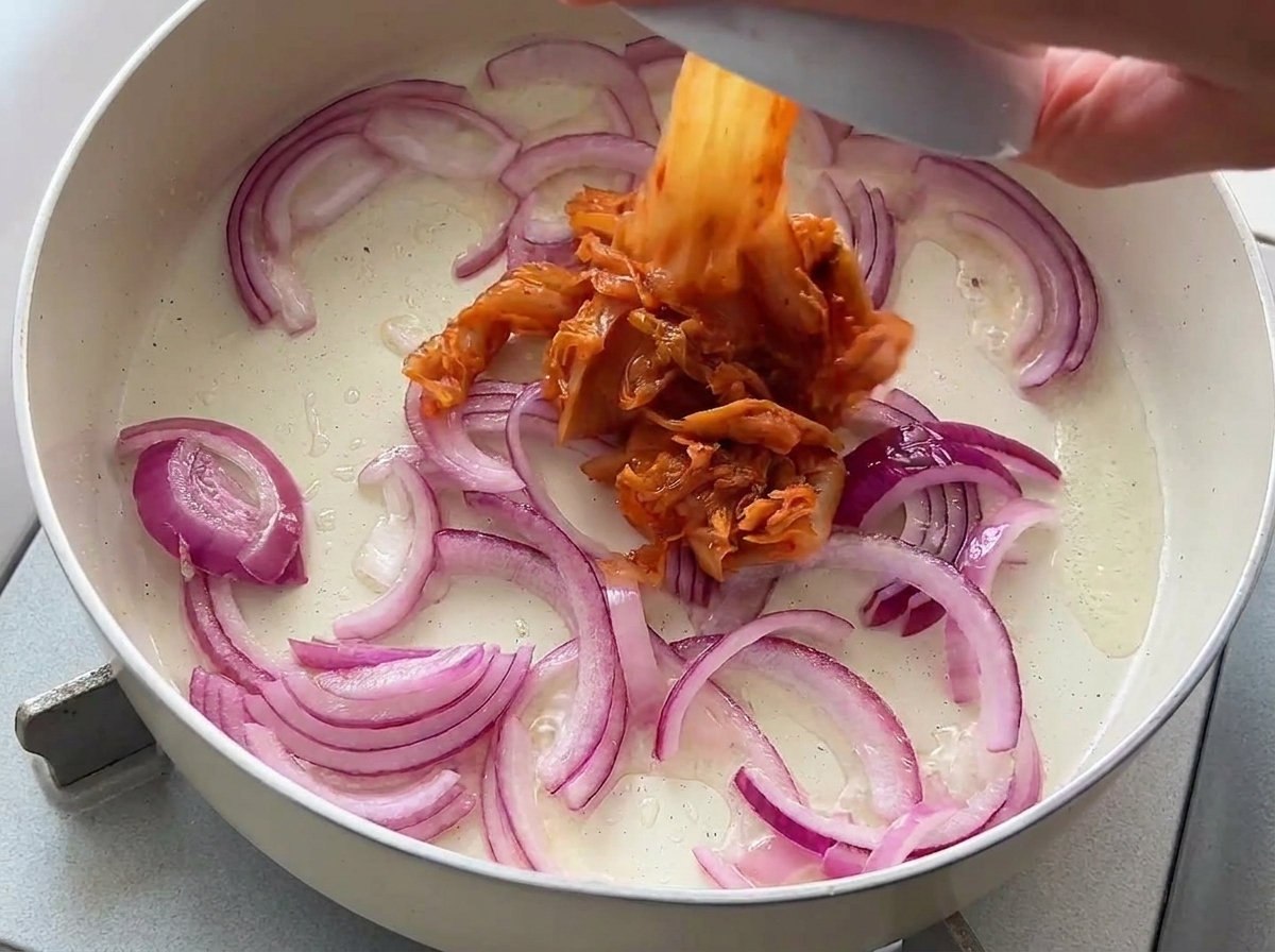 Chopped kimchi being added from a small bowl into a pan of sautéed red onions.