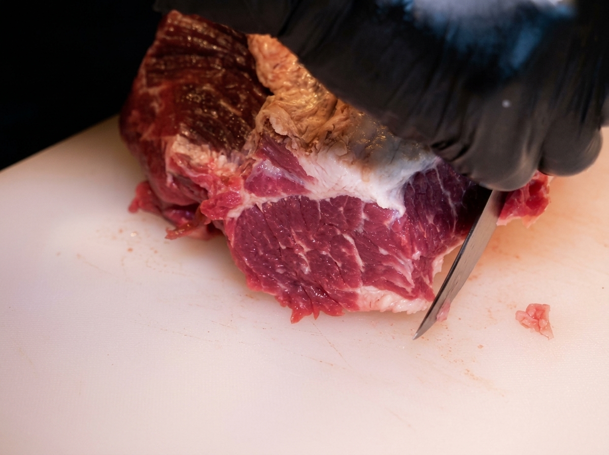 Close up of gloved hands using a knife to trim fat from a large piece of raw beef brisket on a white cutting board