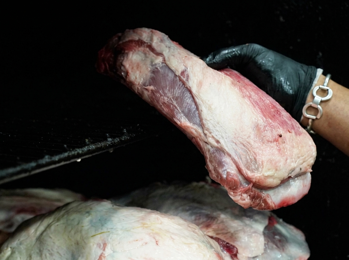 A hand wearing a black glove placing a large cut of raw beef onto the grates of a black smoker.
