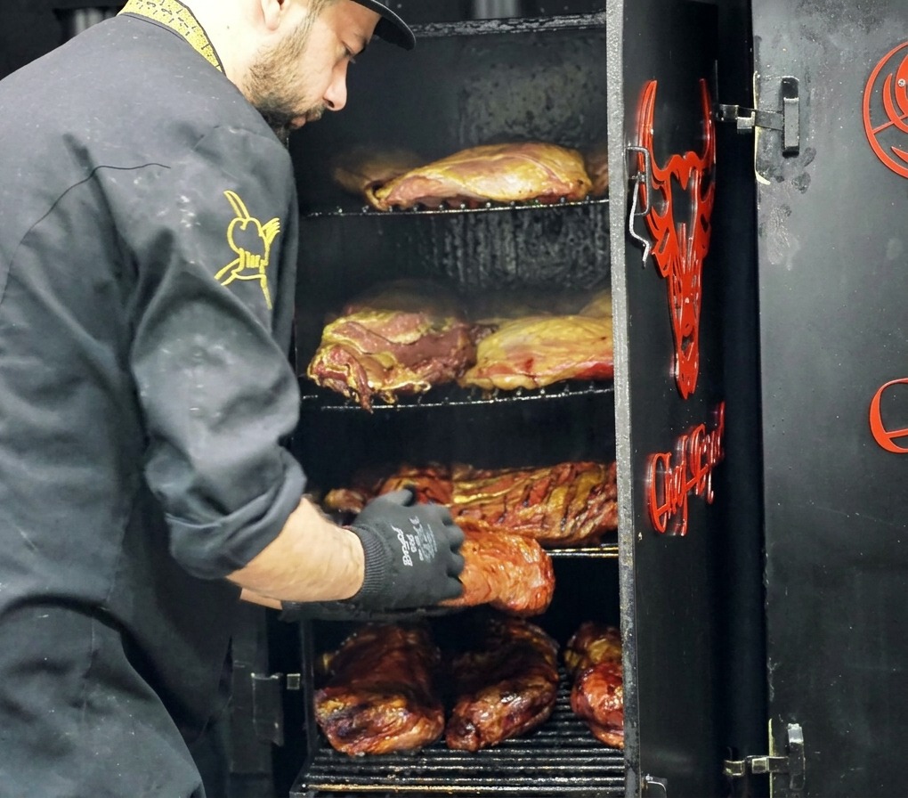 A chef removing a large piece of smoked beef with a dark crust from a multi-rack meat smoker.