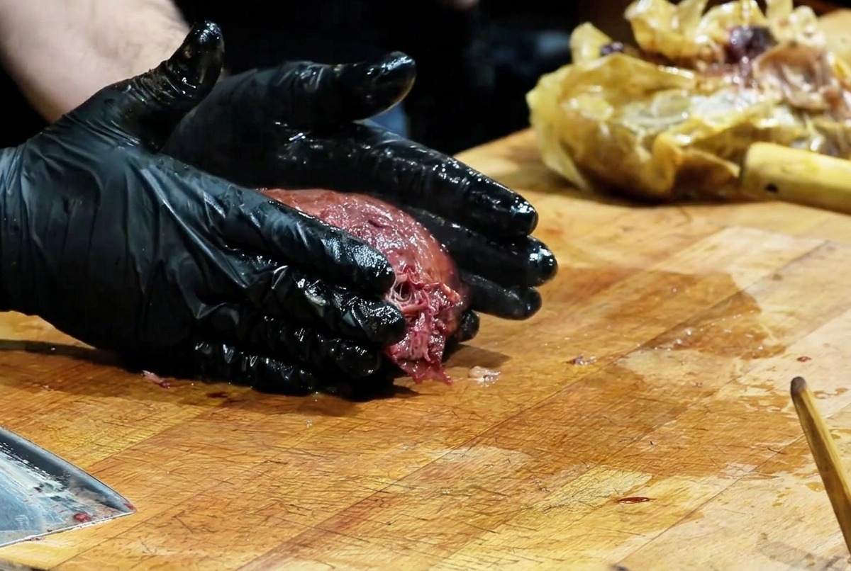 A chef wearing black gloves pressing down on a large dark crusted smoked brisket on a wooden cutting board to shred the meat