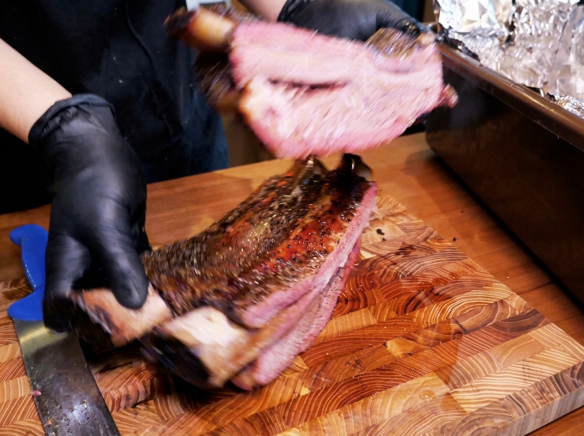 A large rack of smoked beef ribs with a thick dark bark being handled by gloved hands, revealing a juicy pink interior.