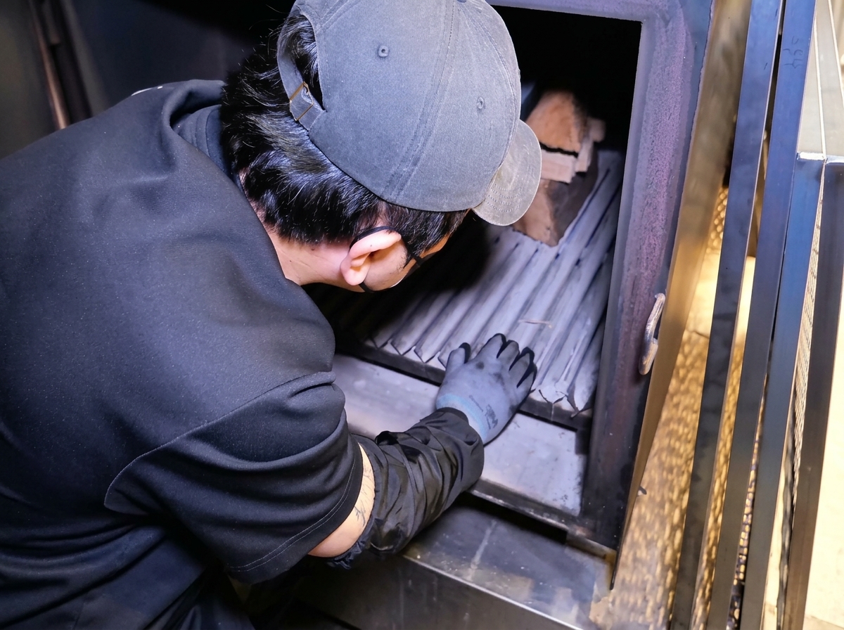 A person wearing a cap and black shirt arranging wood logs inside the firebox of a large metal smoker.