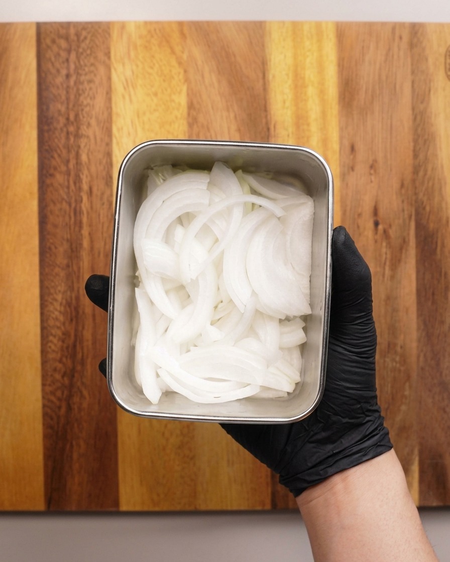 A gloved hand holding a small, square metal container filled with thinly sliced raw white onions.