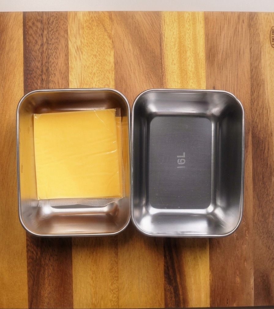 Two small stainless steel rectangular trays on a wooden board, one containing a stack of square yellow cheese slices and the other containing shredded white cheese.