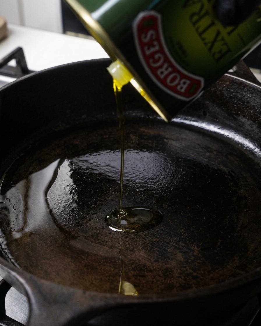 Olive oil being poured from a green tin into a hot, dark cast-iron skillet.