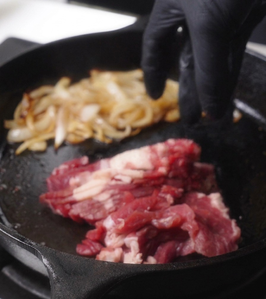 A hand in a black glove adding a pile of thinly sliced raw red beef to a hot cast-iron pan next to a pile of cooked onions.