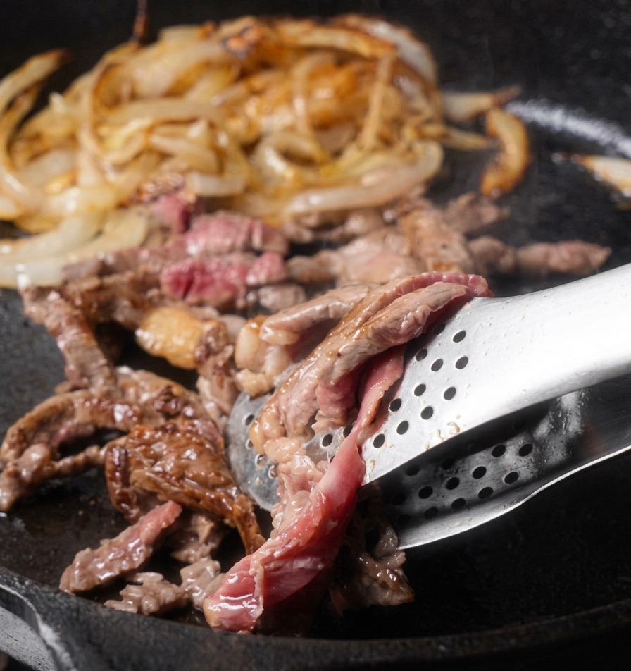 Thin strips of beef being flipped in a cast-iron skillet with metal tongs, showing a mix of browned surfaces and some remaining pink interior.