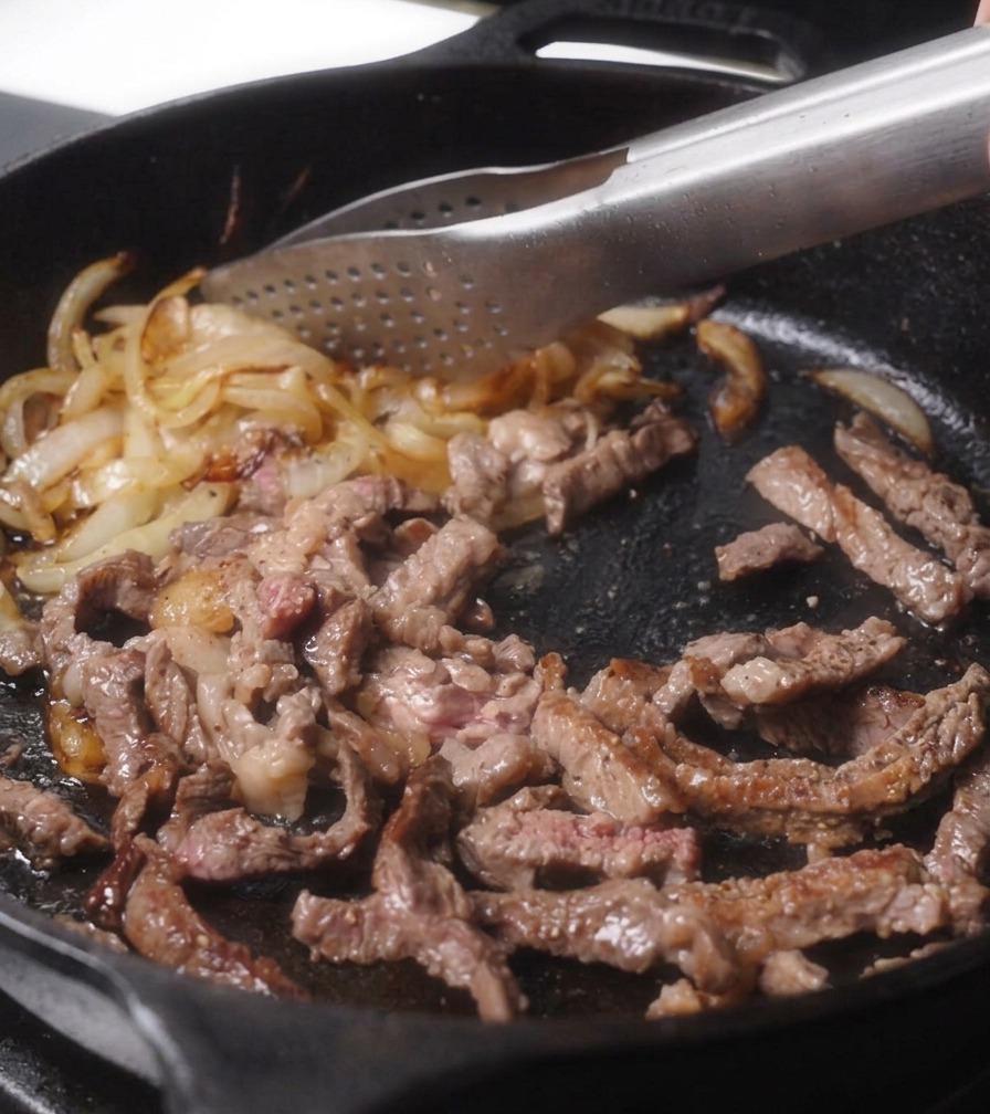 Metal tongs mixing together cooked brown beef strips and golden-brown onions in a cast-iron pan.