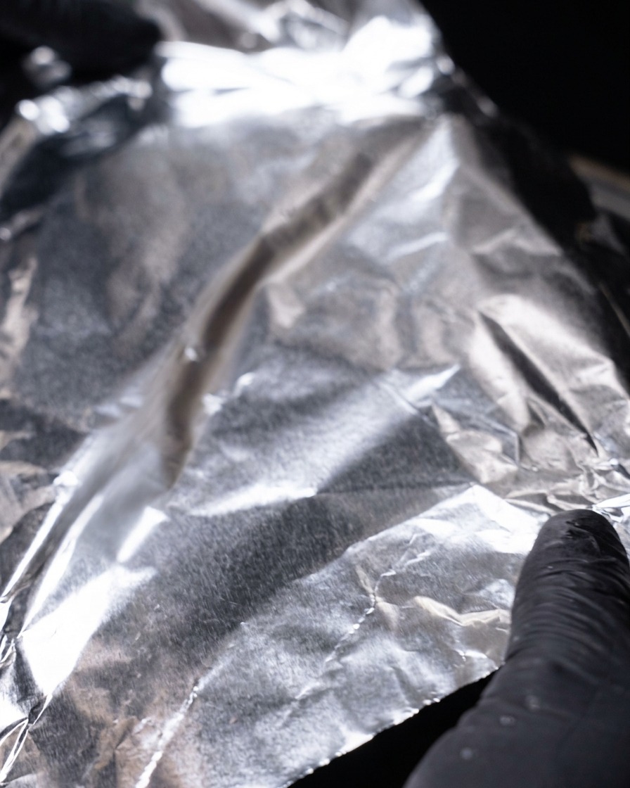 Gloved hands covering a hot skillet with a large sheet of reflective aluminum foil.