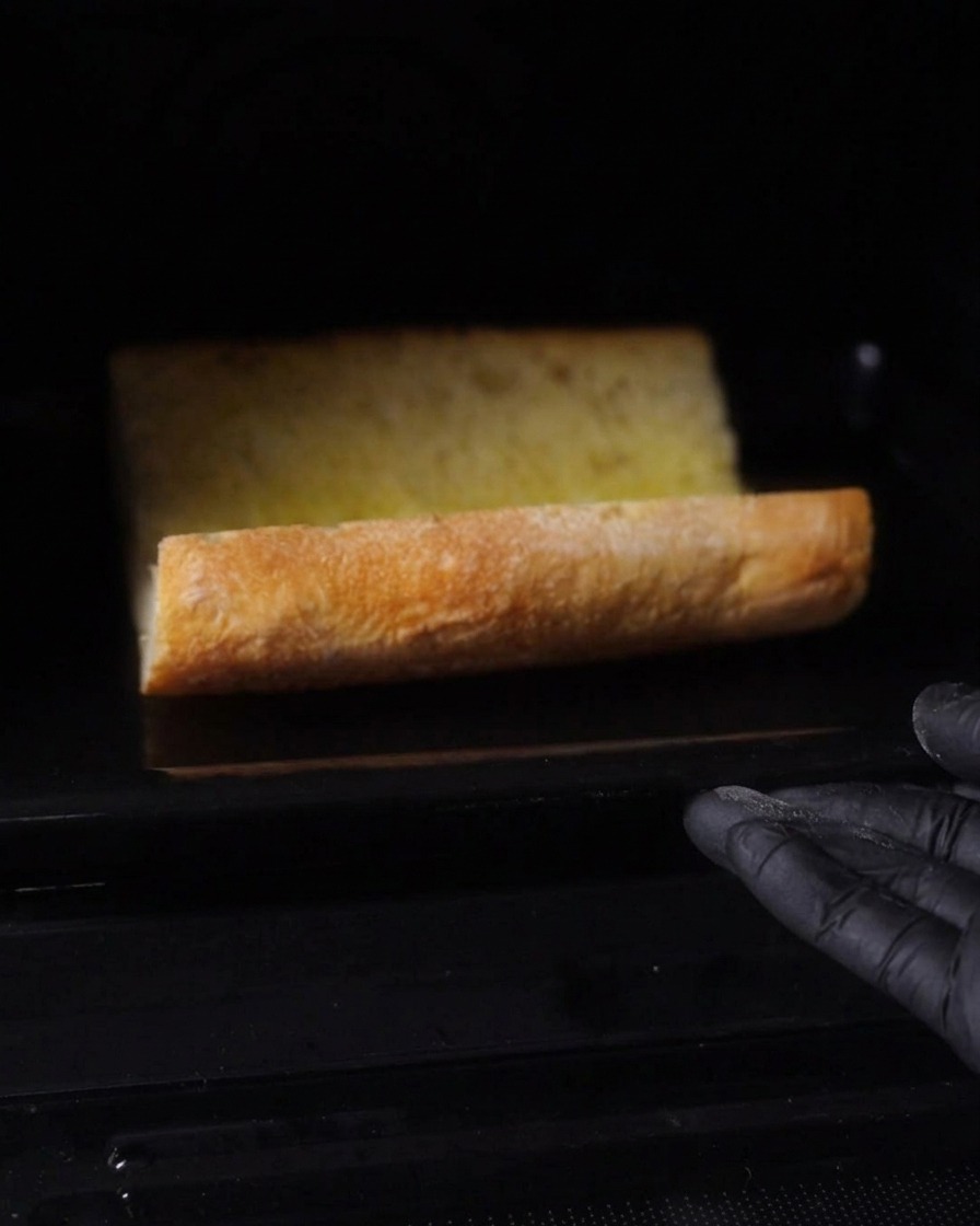 A horizontally split piece of buttered bread baking inside a dark oven.
