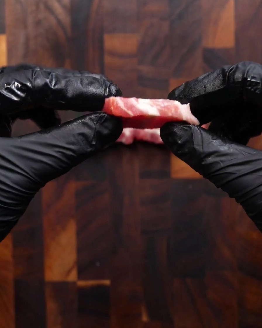 Two hands in black gloves holding up a single, freshly cut slice of raw pork belly to show its coin-like thickness.
