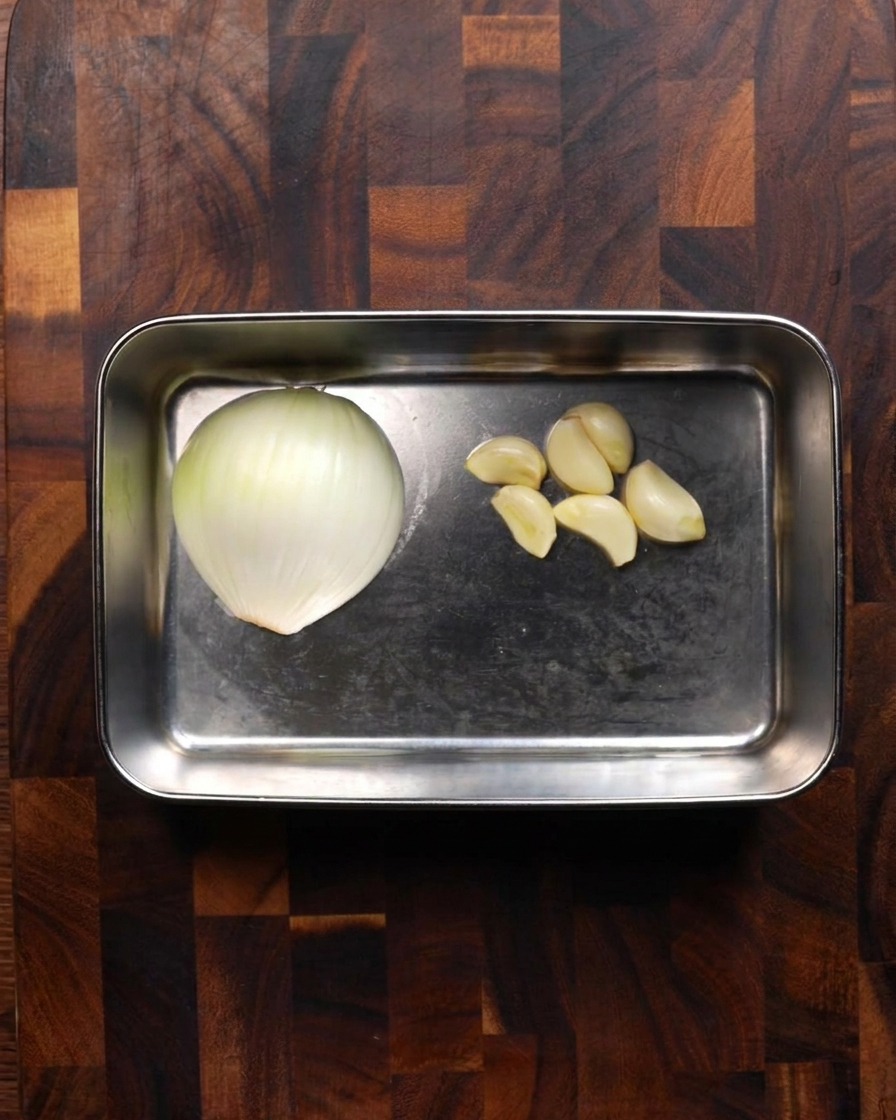 A stainless steel tray holding half of a white onion and several peeled garlic cloves sitting on a wooden board.