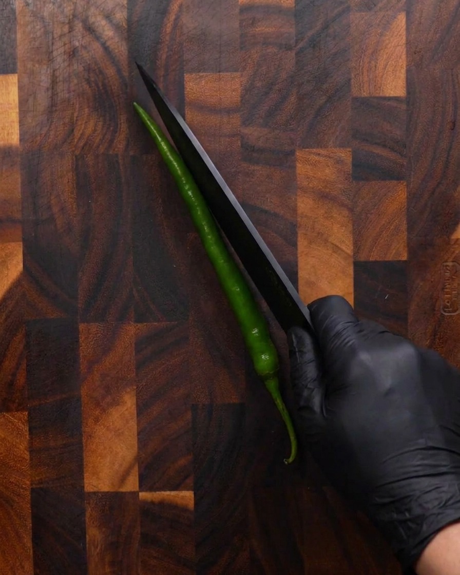 A hand wearing a black glove using a sharp black knife to slice a long green chili pepper diagonally on a wooden cutting board.