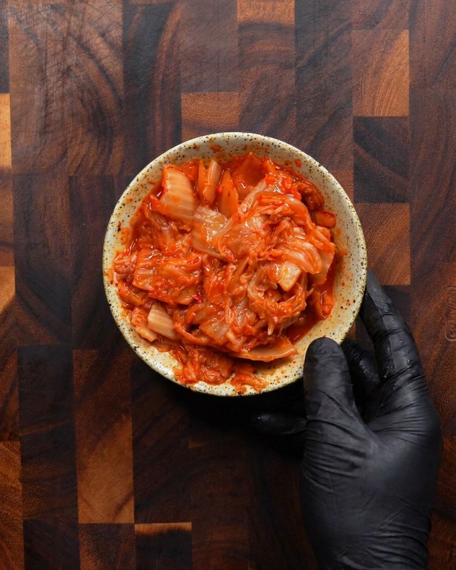 A gloved hand holding a small, rustic speckled bowl filled to the brim with bright red, chopped fermented kimchi.