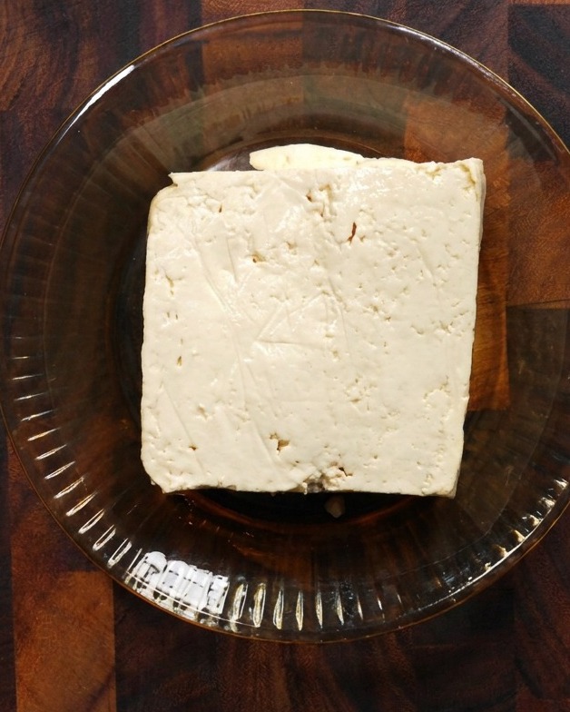 A square block of firm white tofu resting in a shallow glass bowl on a wooden surface.