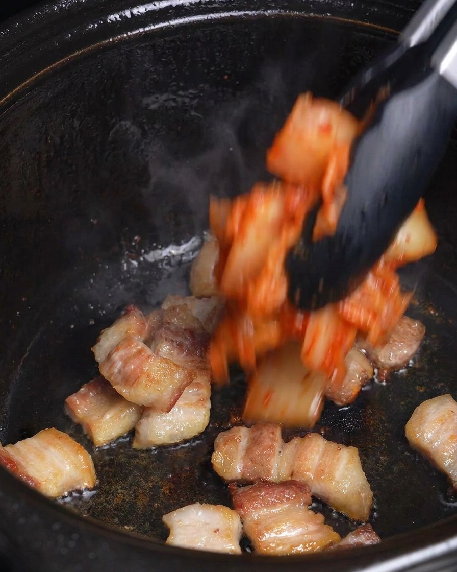 Chopped red kimchi being added to browned pork belly in a black cooking pot with tongs.