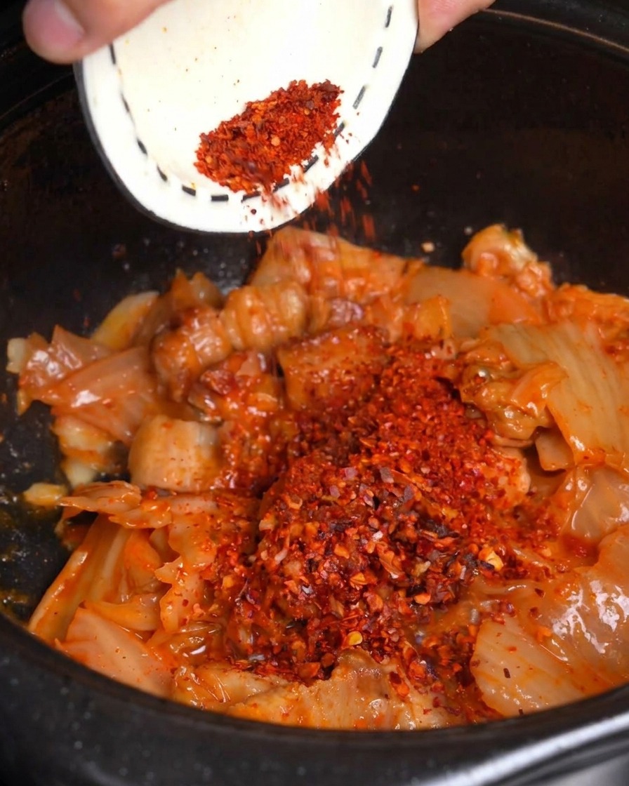 Red chili powder being poured from a small bowl over stir-fried kimchi and pork belly in a black pot.