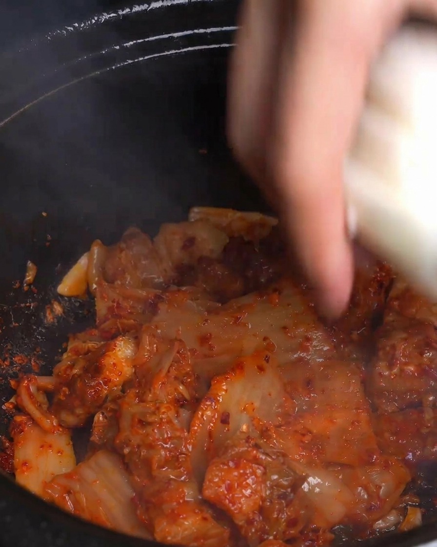 A hand tossing thick slices of fresh white onion into a hot, steaming pot of bright red stir-fried kimchi and pork belly.