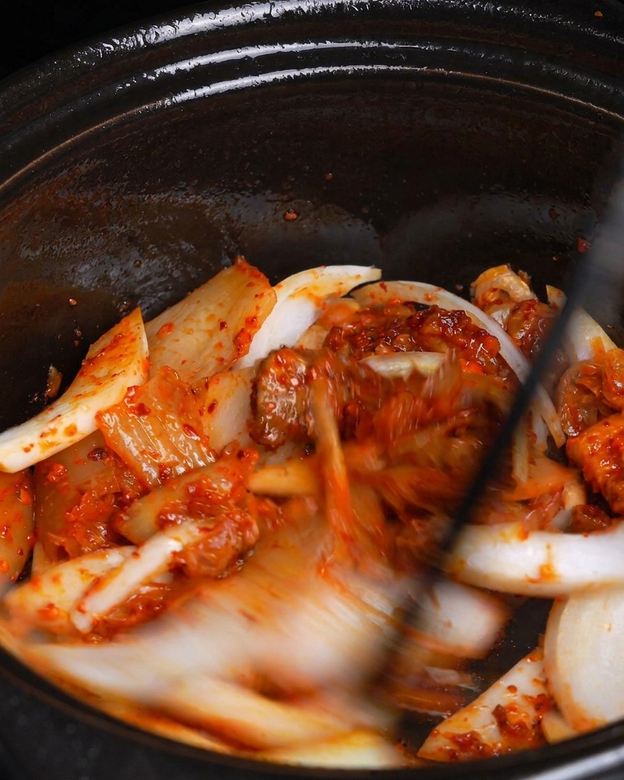 A hand pouring cloudy white liquid from a glass measuring cup into a dark pot filled with cooked pork and red vegetables.