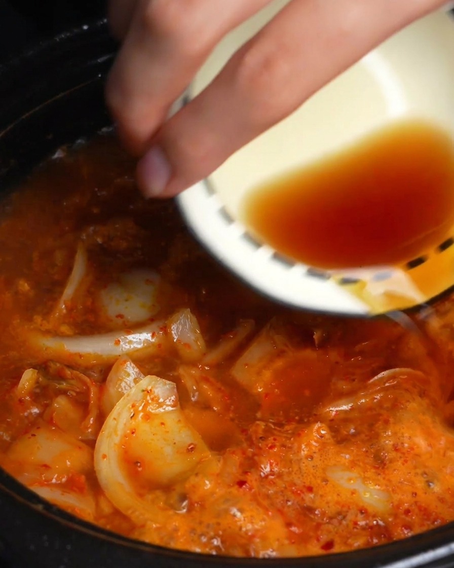 A hand pouring dark soy and fish sauce from a small bowl into a simmering red kimchi broth.