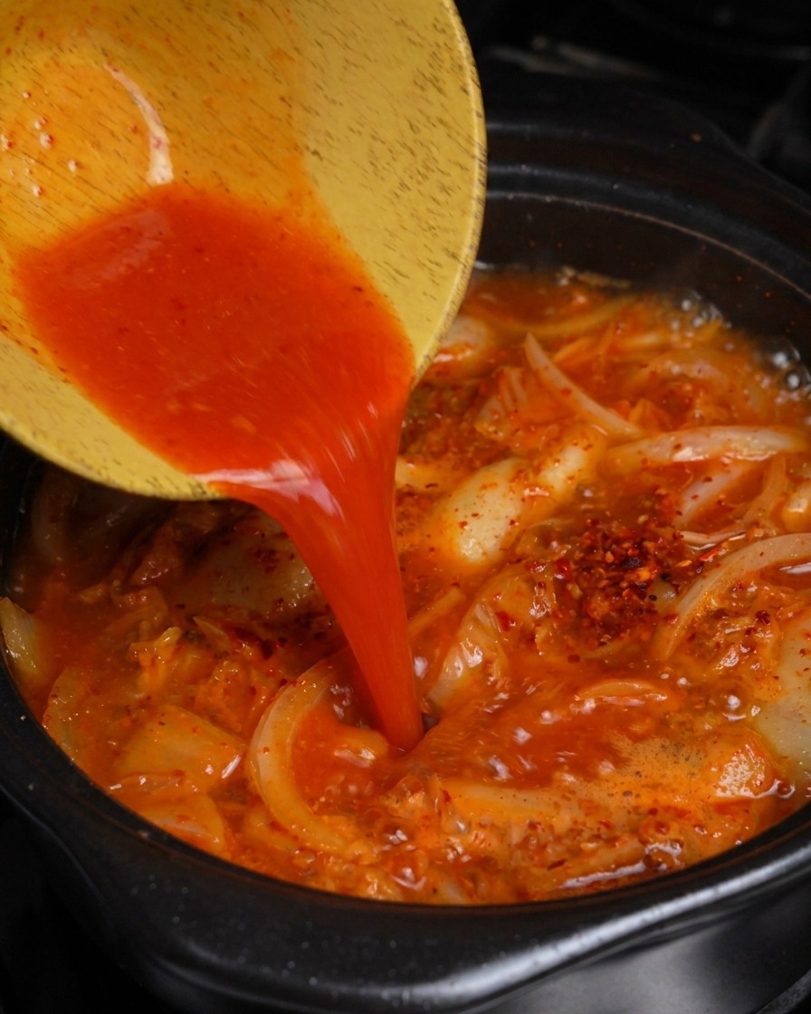 Pouring bright red kimchi juice from a wooden bowl into a pot of bubbling pork and kimchi stew.