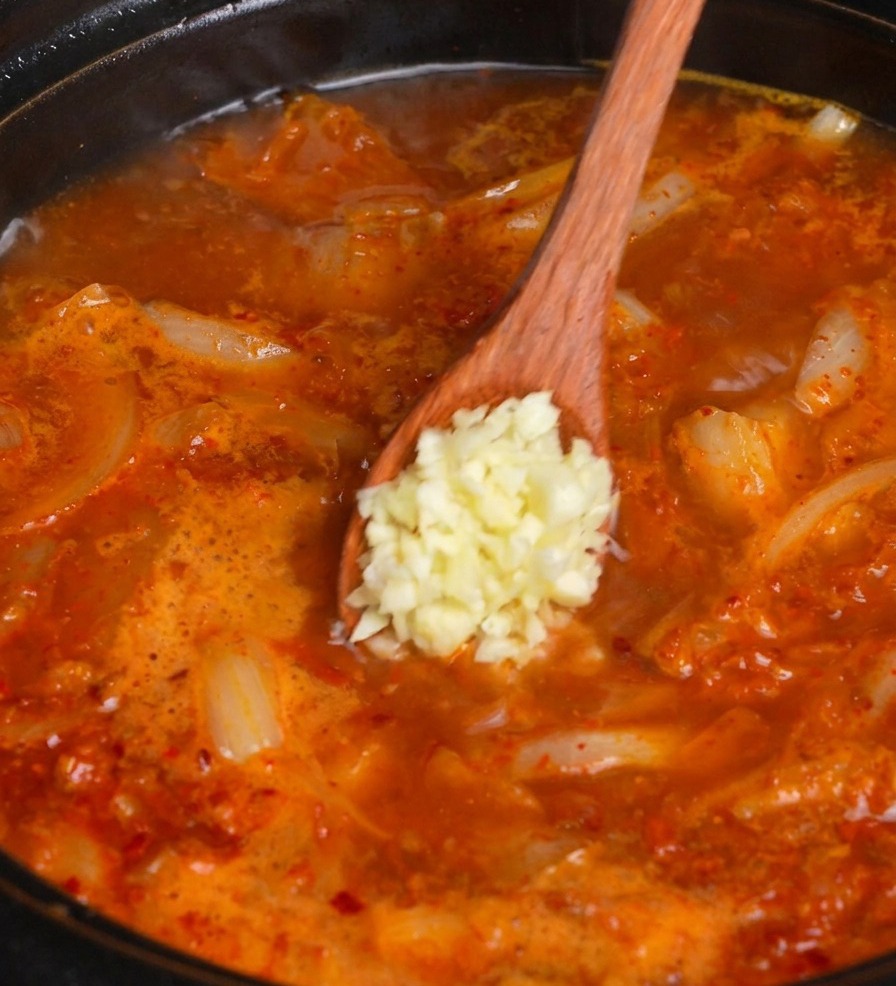 A wooden spoon adding a scoop of fresh minced garlic into a simmering pot of red kimchi soup.