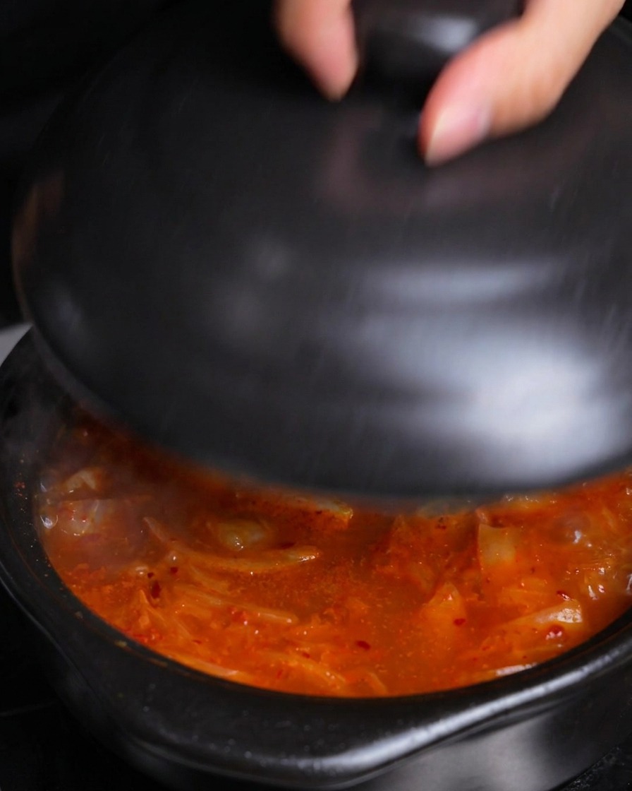A hand placing a black ceramic lid onto a pot of hot, bubbling kimchi stew to let it simmer.