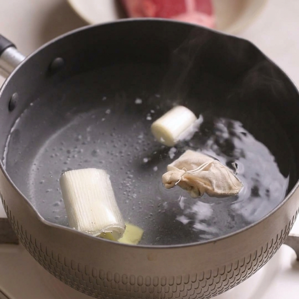 A small pot of boiling water containing chunks of green onion, a slice of ginger, and a small tied spice bag.