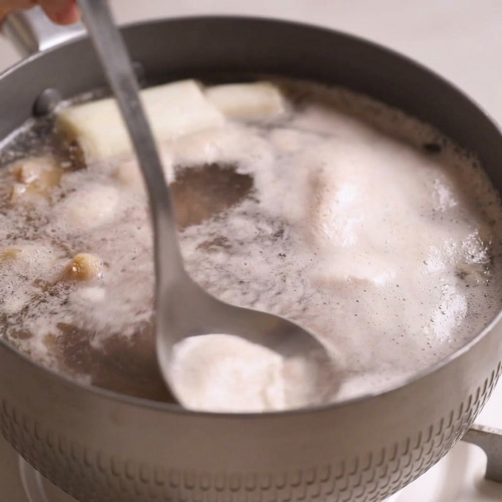 A metal ladle dipping into a pot of boiling beef broth, skimming off grayish foam from the bubbling surface.