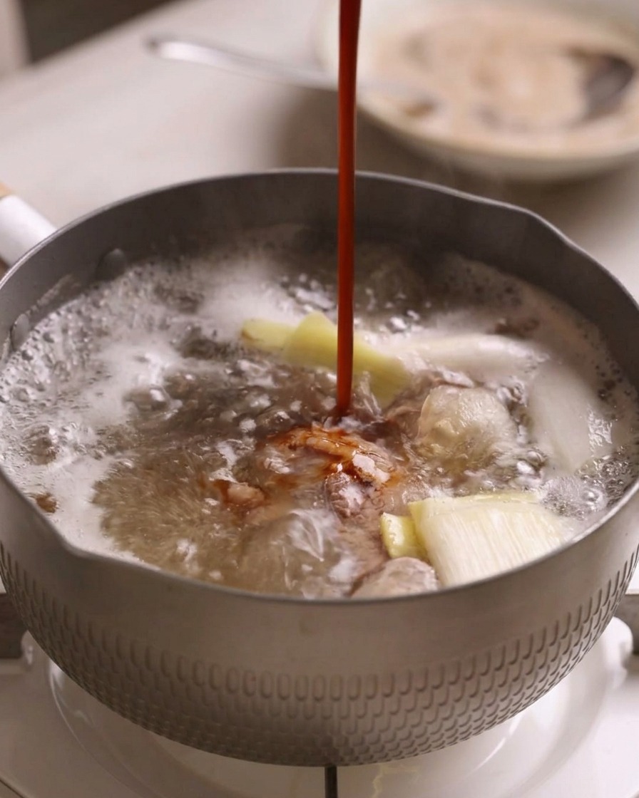 A dark red braising sauce being poured in a steady stream into a pot of boiling beef and aromatics.