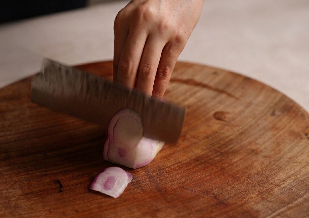 A hand holding a wide cleaver slicing through a half of a red onion on a large, round wooden cutting board.