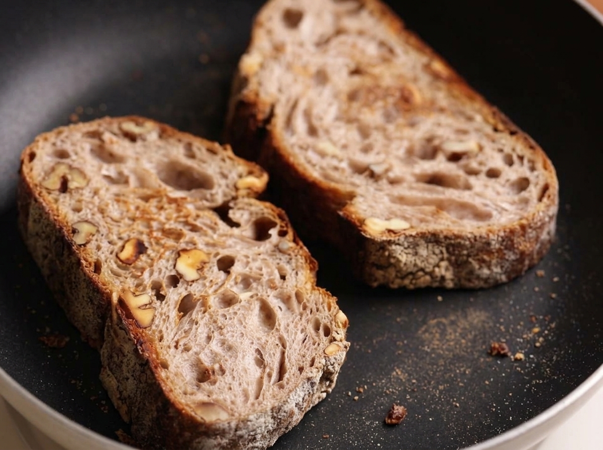 Two slices of rustic artisan bread toasting in a dark, flat skillet.